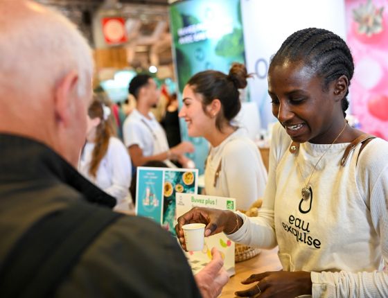 Exposante souriante en interaction avec un visiteur sur son stand à foire de paris, moment de découverte et de dégustation